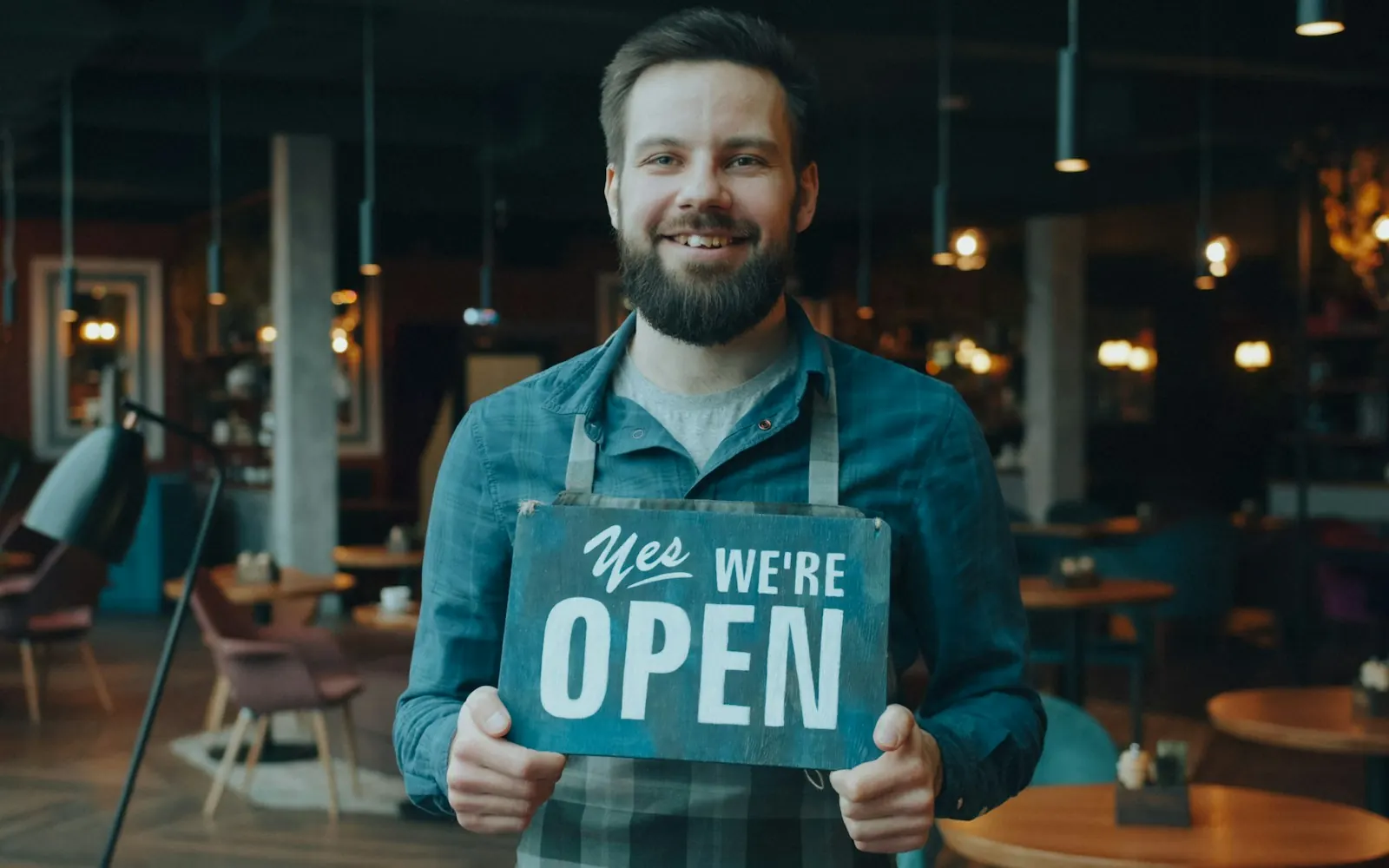 Smiling restaurant owner in an apron holding a sign that reads Yes we're open